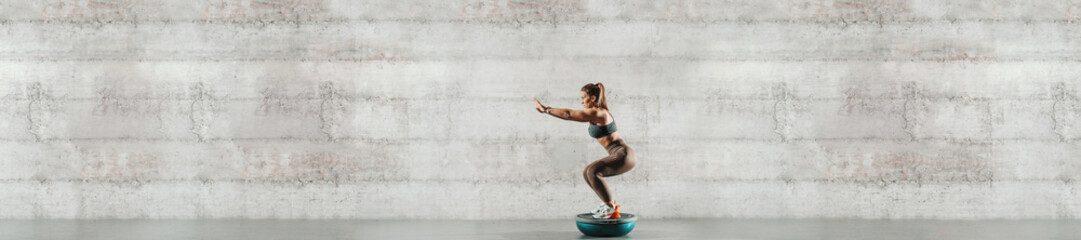 Side view of sporty woman doing exercises on bosu ball. In background gray wall, copy space.