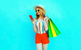 Portrait happy smiling woman holding phone with shopping bags in colorful t-shirt, summer straw hat, sunglasses, red shorts on blue background