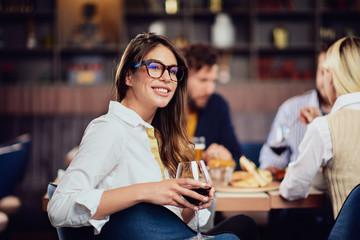 Portrait of gorgeous brunette looking at camera and holding glass with red wine while sitting at restaurat. In background friends eating diner.