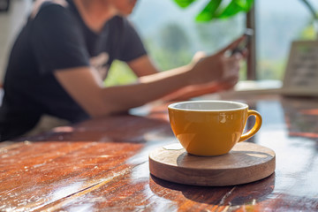 A man in black t-shirt is playing his smartphone near the window with morning sunrise and drinking his hot coffee in yellow cup while waiting for his friends in a cafe . 