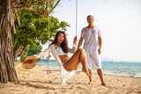 Multinational couple relaxing on the beach in Thailand. The woman sit on swings, man behind
