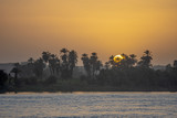The Egyptian sun rises behind palm trees on the banks of the Upper Nile River in the Aswan Governorate, Egypt.