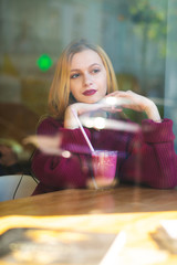 Beautiful young model spending time in the restaurant with a cup of coffee. Shot through window glass