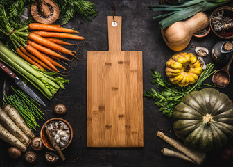 Autumn seasonal food background with colorful various pumpkins and organic farm vegetables around wooden cutting board, top view. Healthy vegetarian cooking. Thanksgiving or Halloween recipes.