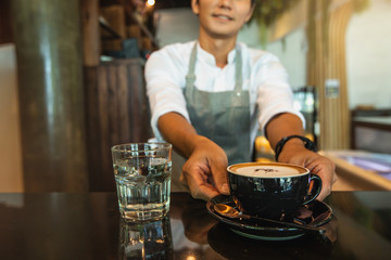 Portrait of smiling barista with coffee cup in front of customers at coffee shop. Portrait of happy young male coffee shop owner standing with barista working behind the counter bar.