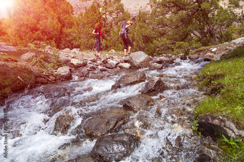 Poster Two hikers crossing fast flowing river People going across mountain creek with f