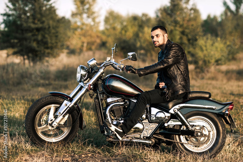 young brutal man in a black jacket and glasses sits near a motorcycle Slika na platnu