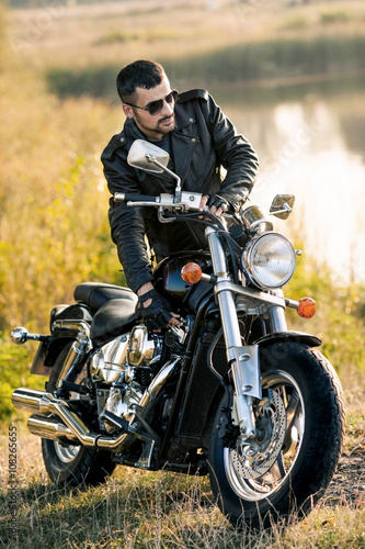 young brutal man in a black jacket and glasses standing near a motorcycle Slika na platnu
