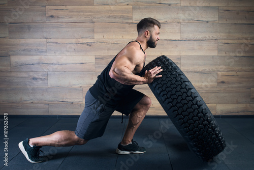 Poster Muscular man with tattoos and beard pushing a tire in a black tank top and grey