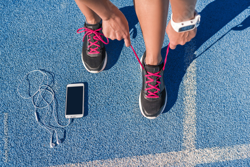 mujer-del-corredor-que-ata-los-cordones-de-los-zapatos-para-correr-preparandose-para-la-carrera-en-la-pista-de-atletismo-con-telefono-inteligente-y-auriculares-para-escuchar-musica-en-el-telefono-movil-atleta-preparandose-para-el-entrenamiento-cardiovascular-pies-en-el-suelo