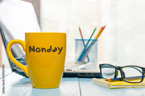Monday written on yellow coffee or tea cup at wooden boards table, workplace, office sunlight morning background