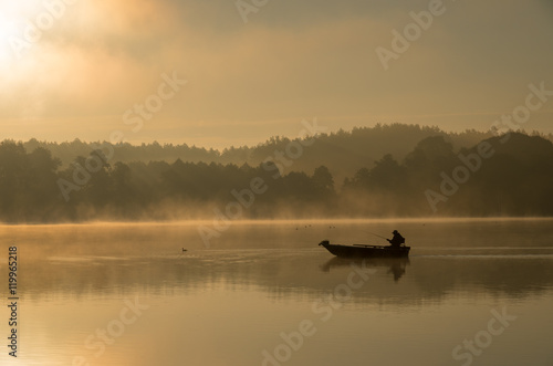 Obraz Fotograficzny Człowiek wędkujący na łódce mglisty, złoty poranek.