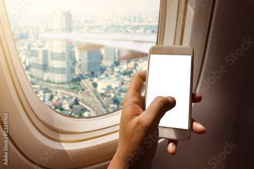 Asian man hand holding blank screen smart-phone on board of airplane near window seat and wing