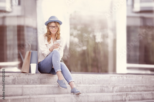 Time to communicate / Happy young woman with a disposable coffee cup and shopping bags sitting on the stairs and using her smartphone for communication via wi-fi internet in a summer park.