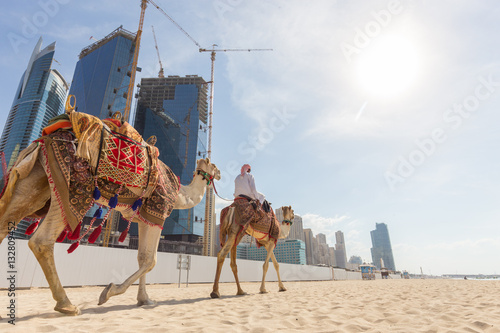 Tour guide offering tourist camel ride on Jumeirah beach on in Dubai, United Arab Emirates. Luxury Dubai Marina skyscrapers in background.