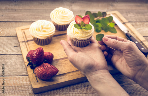 Poster Female hands decorate cupcakes with fresh strawberry and leaves of mint on a wooden background