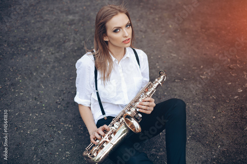 Obraz na plátně Young attractive girl in white shirt with a saxophone sitting sits on the earth - outdoor