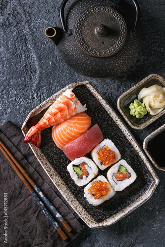 Sushi Set nigiri and sushi rolls in dark ceramic plate with soy sauce, iron teapot and chopsticks over black stone texture background. Top view with space. Japan menu
