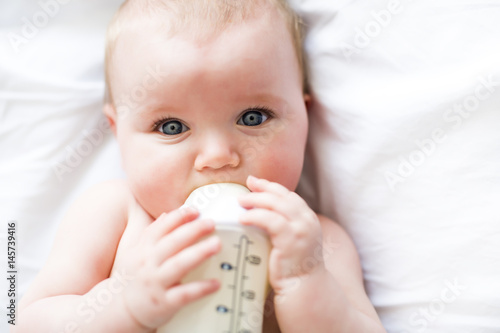 Pretty baby girl drinks water from bottle lying on bed. Child weared diaper in nursery room.