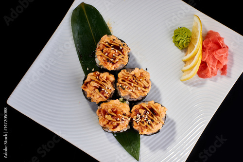 Baked Sushi Roll with crab, eel fish and spicy salmon served with vasabi, pickled ginger and lemon slice on white plate on black background