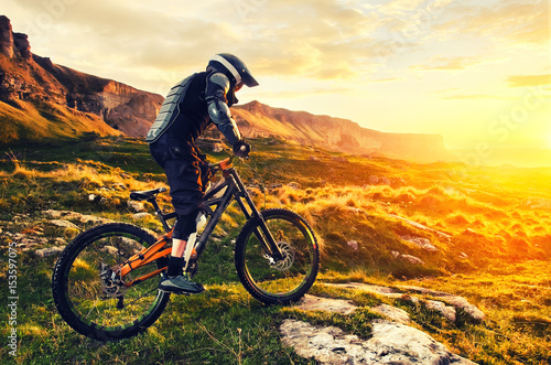 Plakat foto The rider in full protective equipment on the mtb bike is riding toward the sunset in the rays of the sunset sun against the background of the rocks of the setting sun and clouds
