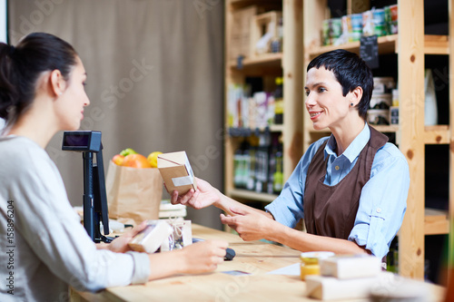Helpful middle-aged shop assistant giving advice to customer while standing at counter in small store with organic food