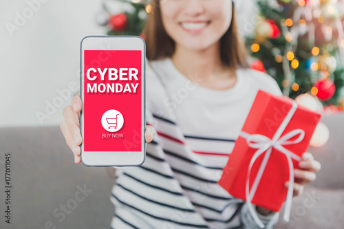 Young beautiful Asian Woman hands holding gift box and smartphone with cyber Monday screen.Smile face in room with Christmas tree decoration for holiday background.