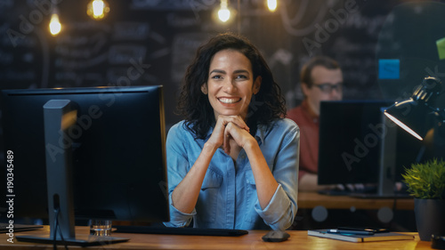 Beautiful Female Office Employee Works at Her Desk on a Personal Computer, She Interrupts Her Work and Smiles Charmingly for the Camera. In the Background Coworker in the Creative Office.