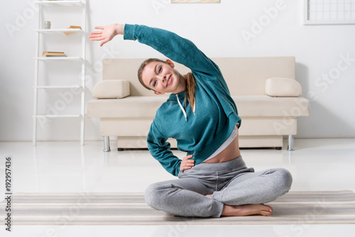 jeune-fille-souriante-assise-en-position-du-lotus-sur-tapis-de-yoga-et-etirement-a-la-maison