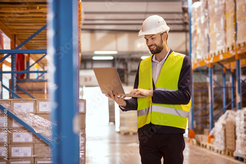 Modern device. Happy cheerful man holding a laptop while using in for doing the inventory in the warehouse