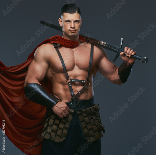 Portrait of ancient Greece warrior with a muscular body dressed in battle uniforms holds sword on his shoulder.