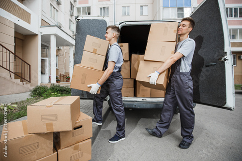 Two young handsome smiling workers wearing uniforms are unloading the van full of boxes. The block of flats is in the background. House move, mover service. © Leika production
