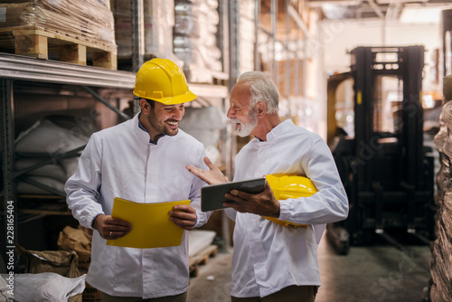 Team work. Picture of two man warehouse employes smiling and looking together at digital tablet. Older college is learning younger one how to solve a problem.Standing in big warehouse.