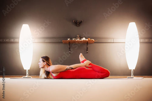 Side view of attractive woman in a red tight-fitting gym suit doing dhanurasana lying on rug on floor. Concept of spinal flexibility extending youth and prevention of diseases of the abdominal cavity © Nadezhda
