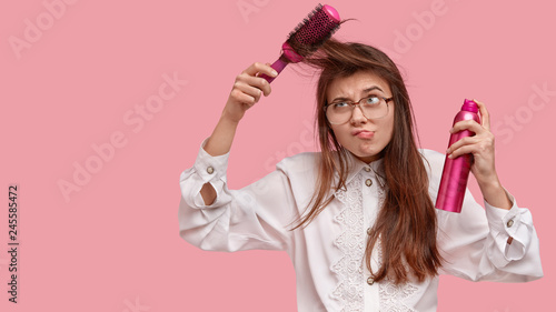 Dissatisfied young woman brushes hair, uses comb, frowns face, holds hairspray, wears white old fashionable blouse, prepares for first date, isolated over pink background with copy space aside © Wayhome Studio