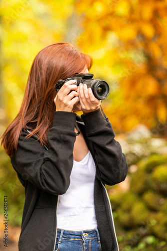 A young woman with reddish hair takes pictures with a reflex camera in a forest