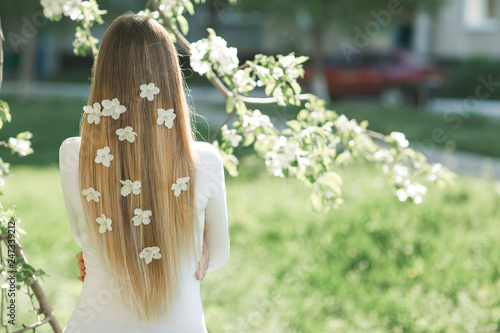Unrecognizable woman standing back side to camera with long blond hair with flowers in her hair. Female on spring background. Lady outdoors. © olenachukhil
