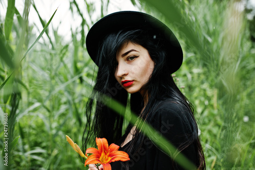Sensual girl all in black, red lips and hat. Goth dramatic woman hold orange lily flower on common reed. © AS Photo Project
