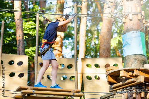 Cute young boy in helmet with climbing equipment in the rope amusement park. Summer camp, holidays concept