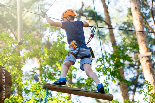 Cute young boy in helmet with climbing equipment in the rope amusement park. Summer camp, holidays concept