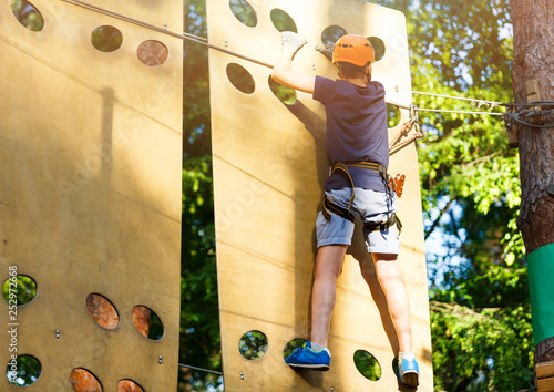 Cute young boy in helmet with climbing equipment in the rope amusement park. Summer camp, holidays concept