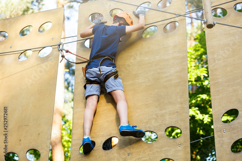 Cute young boy in helmet with climbing equipment in the rope amusement park. Summer camp, holidays concept