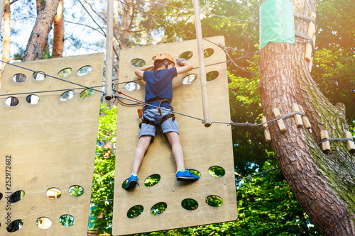 Cute young boy in helmet with climbing equipment in the rope amusement park. Summer camp, holidays concept