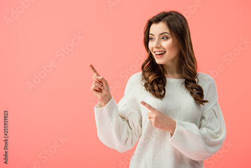 smiling and beautiful woman in white sweater pointing with fingers and looking away isolated on pink © LIGHTFIELD STUDIOS