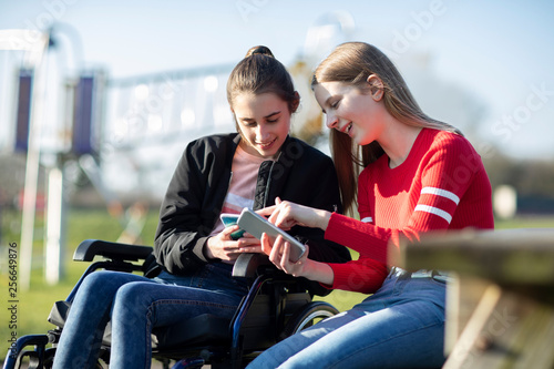 Teenage Girl In Wheelchair Looking At Mobile Phone With Friend In Park © Daisy Daisy