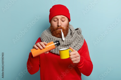 Sad bearded red haired young man measures temperature, pours tea from thermos bottle, wears warm winter clothes, being fed up of seasonal disease, isolated on blue background. Illness concept