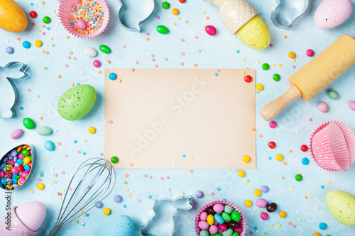 Easter baking background with paper card, rolling pin, whisk, decorative eggs, cookie cutters, candy and colorful confetti on kitchen table top view. Flat lay. © juliasudnitskaya