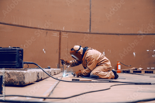 Worker in protective suit and mask crouching and welding in metal tower at construction site. © dusanpetkovic1