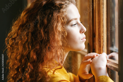 Teen curly ginger girl in the yellow shirt looking to the big window at the loft interior © proimagecontent