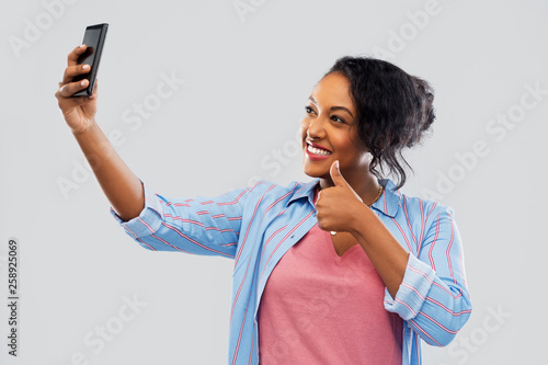 technology and gesture people concept - happy african american woman taking selfie by smartphone and showing thumbs up over grey background © Syda Productions